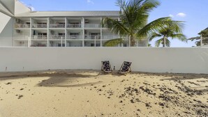 Un hôtel situé en bord de mer, avec une façade blanche, de nombreux balcons et deux chaises longues sur la plage de sable.