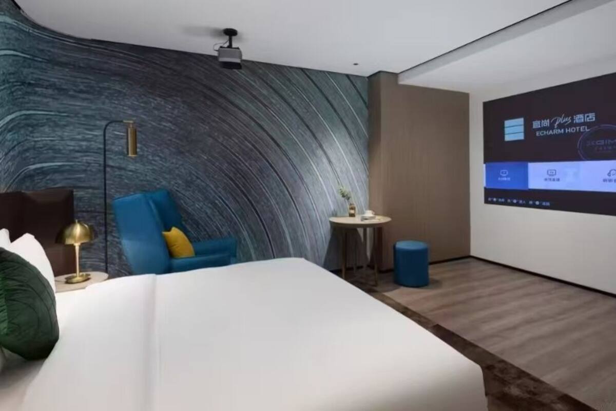 Una camera d'albergo moderna con parete curva, un letto, una poltrona blu, un piccolo tavolino rotondo e uno schermo di proiezione che mostra informazioni dell'hotel.