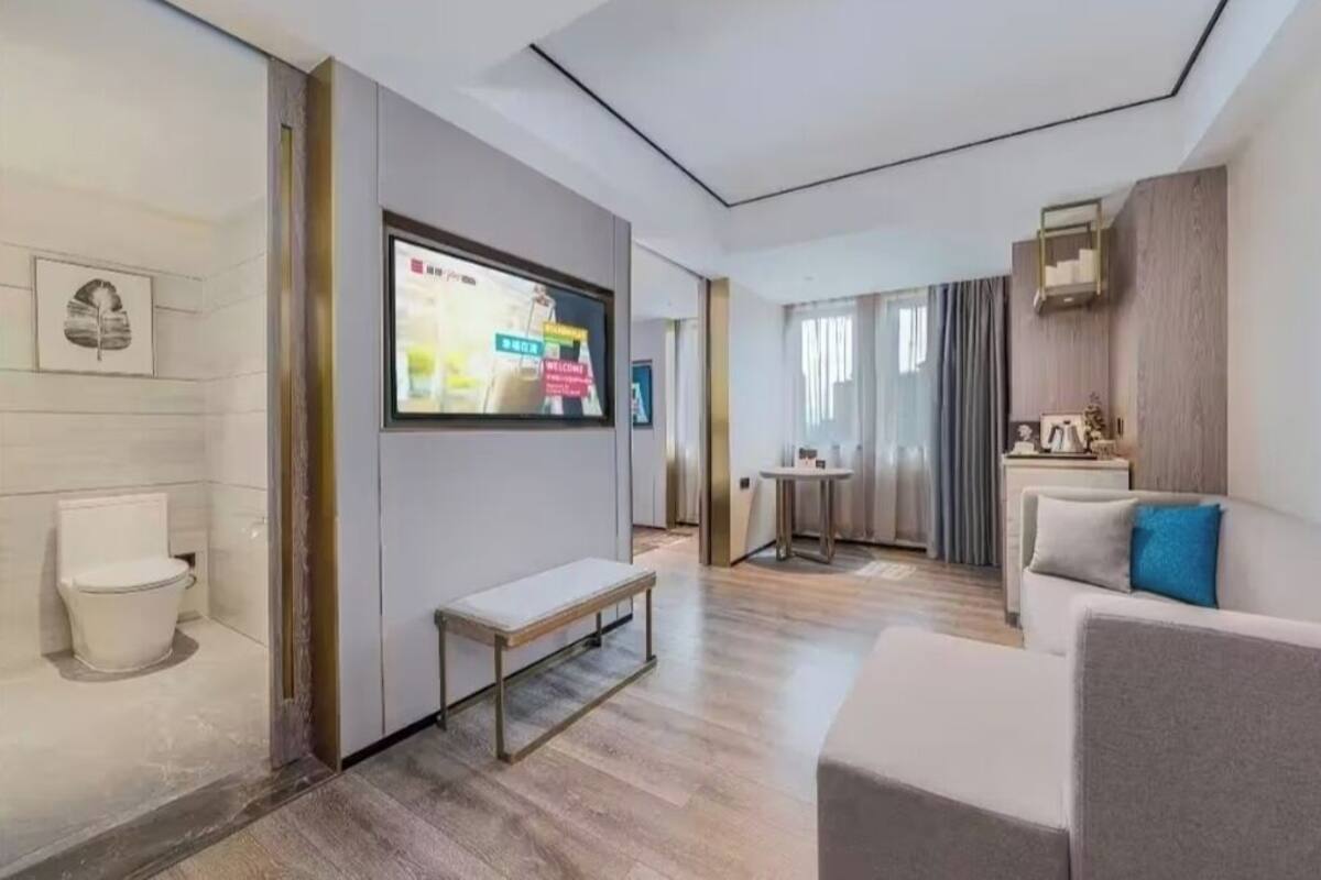Una camera d'albergo moderna con pavimento in legno, un divano bianco, una panca, una TV a parete e un tavolino con una lampada.