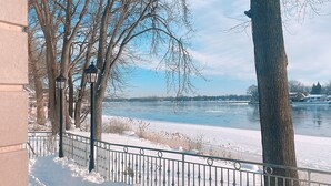 A snow-covered riverbank with a metal fence, bare trees, and a lamp post. - Luxury Riverside Villa with Magnificent RiverViews (Laval)