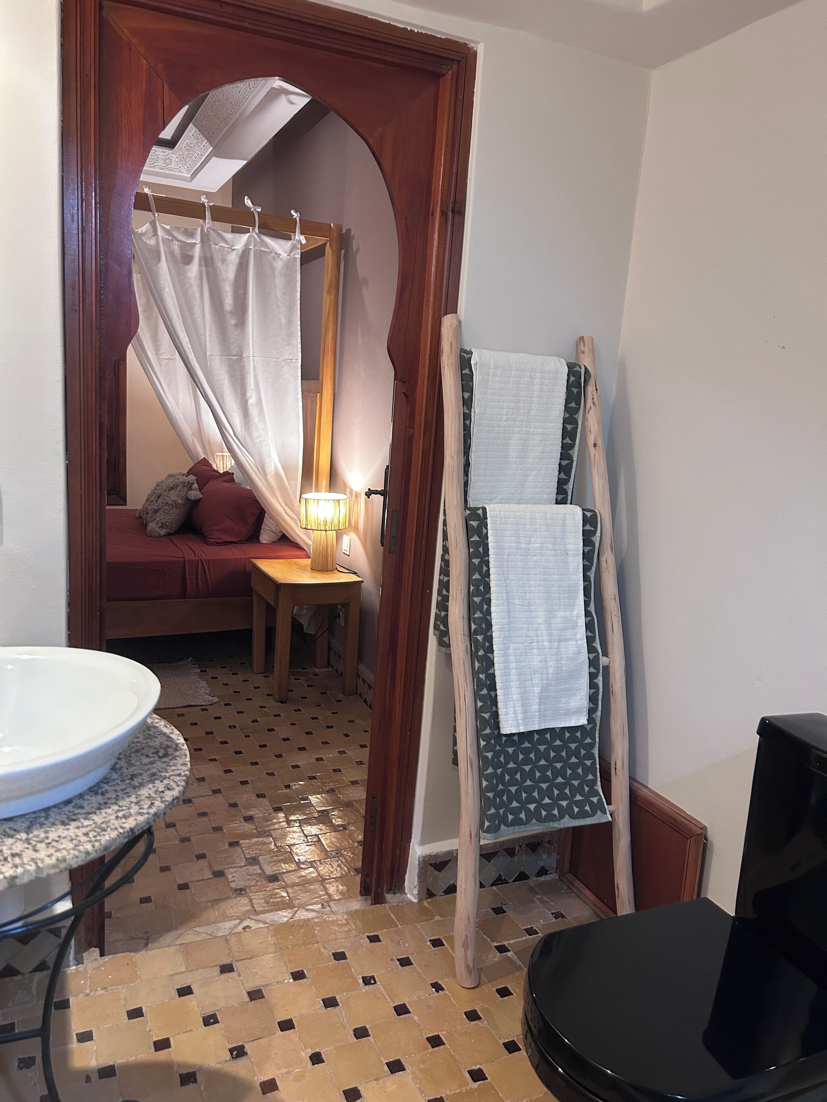 A bathroom with a sink, a mirror, a wooden door, a tiled floor, and a small table with a lamp.