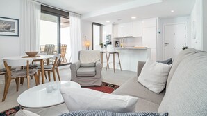 Ein modernes Wohnzimmer mit einer Couch, Sesseln und einem Esstisch. Es gibt eine Küchenzeile mit weißen Schränken und einem Esstisch.