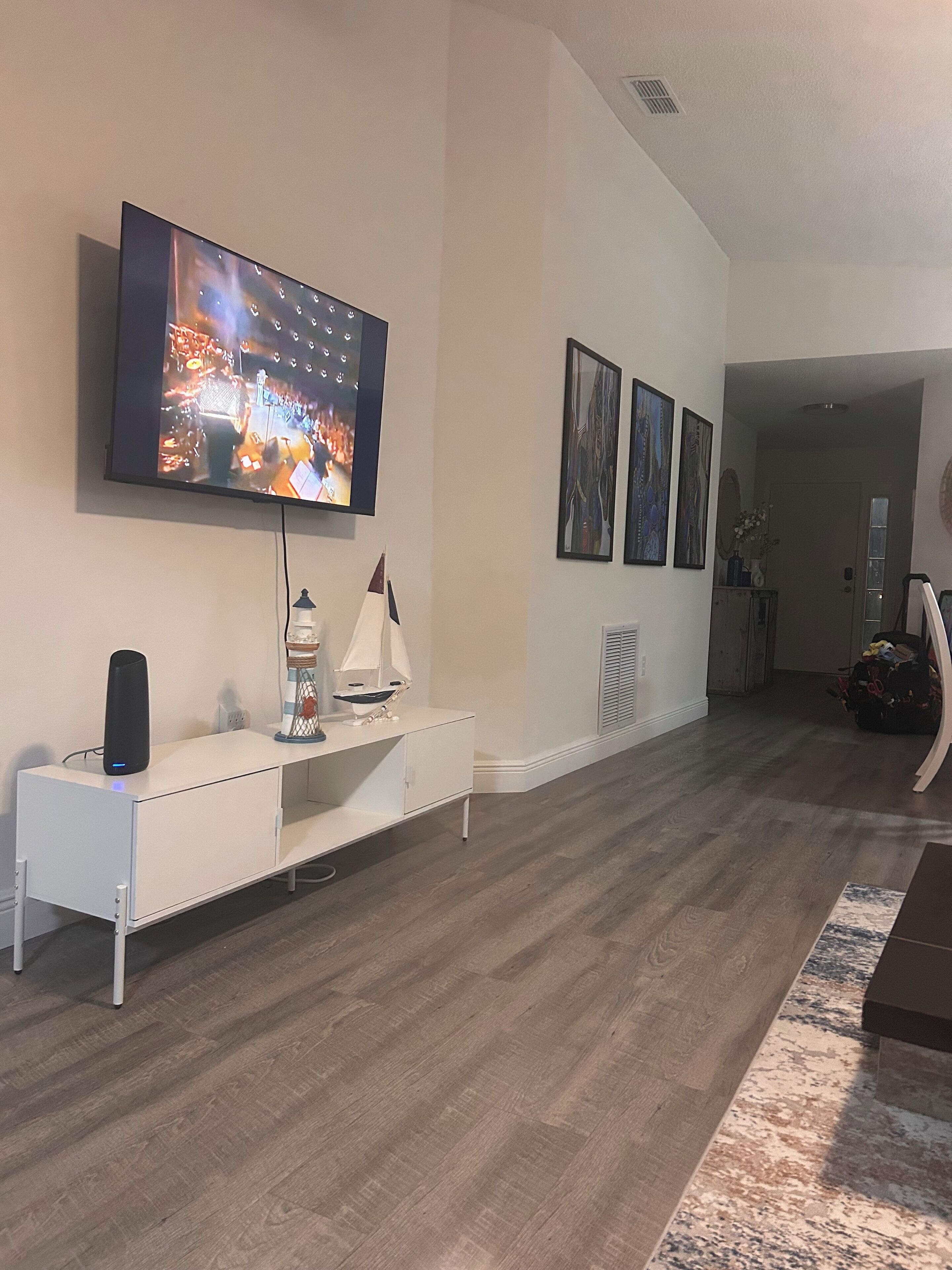 Una sala de estar con un televisor de pantalla plana montado en la pared, un mueble bajo blanco, un modelo de velero pequeño y una alfombra estampada.