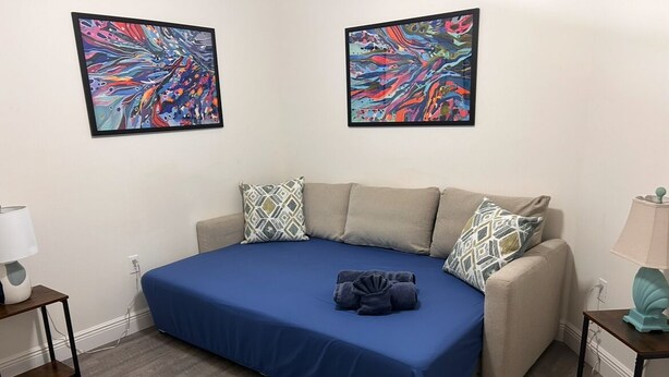 A bedroom with a bed, two pillows, a blue bedspread, a nightstand, a lamp, and a wall-mounted artwork.