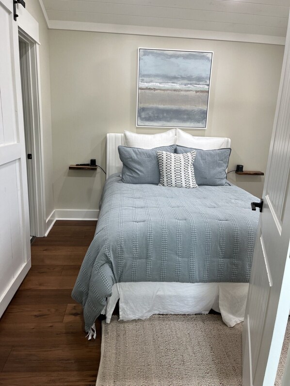 A bedroom with a bed, a bedside table, a nightstand, a wall-mounted artwork, and a wooden floor. - Romantic Getaway at Honeymoon Cottage (Pisgah)