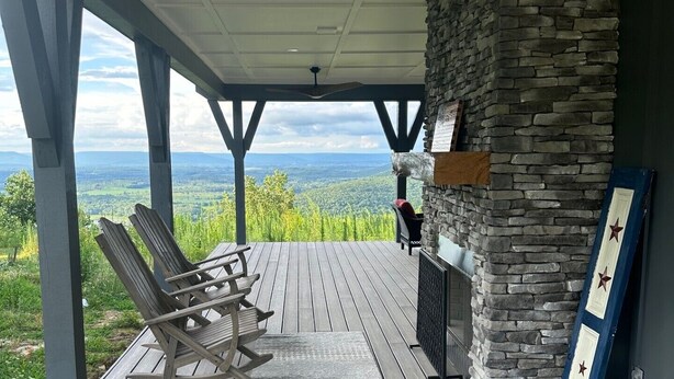 A covered outdoor deck with a stone fireplace, wooden chairs, and a view of the landscape. - Romantic Getaway at Honeymoon Cottage (Pisgah)