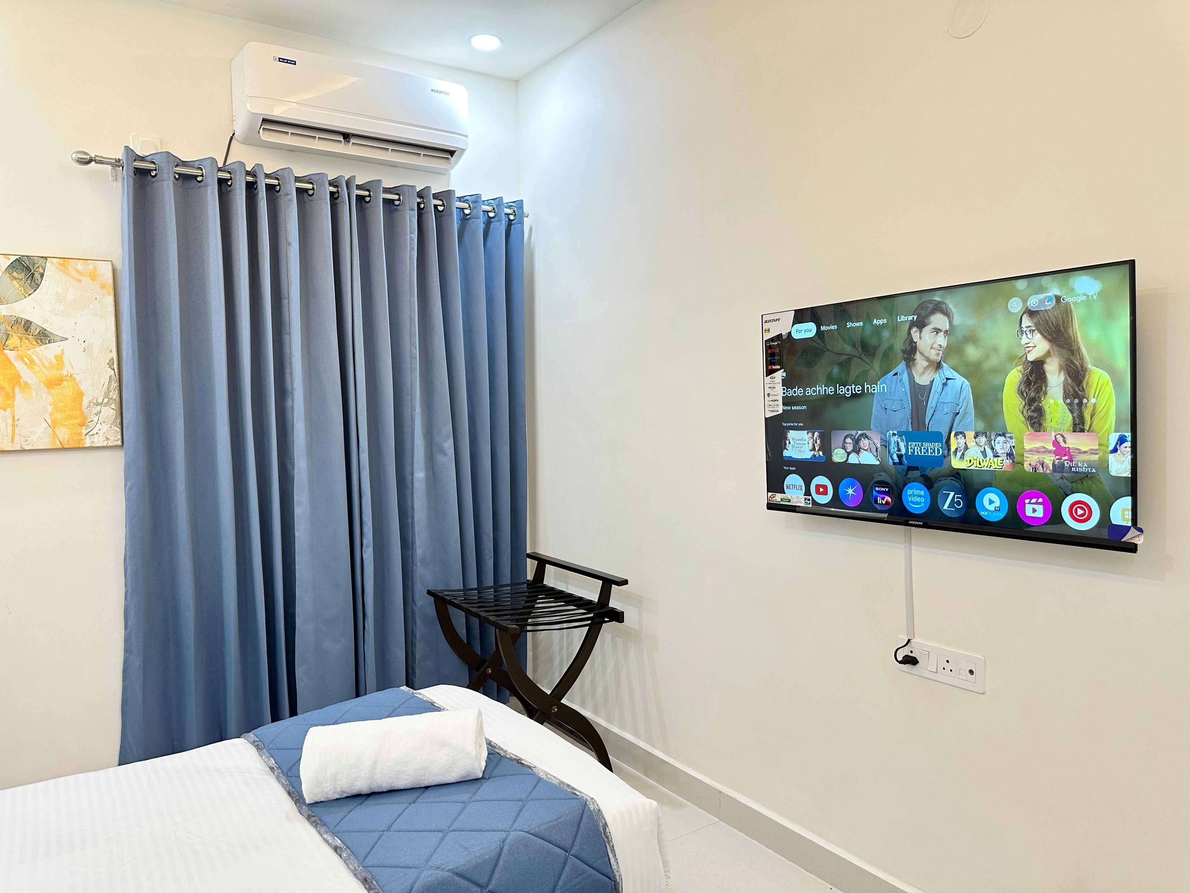 A hotel room with a bed, a TV mounted on the wall displaying a streaming service interface, a ceiling air conditioner, and a window with curtains.
