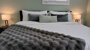 A neatly made bed with a fluffy grey blanket, two bedside lamps, and a window with blinds.