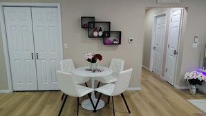 A dining area with a round table and four white chairs, a vase with flowers, and a wall-mounted shelf with decorative items.