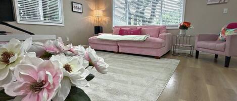 Una sala de estar con un sofá rosa, una mesa de centro, un tapete y flores artificiales de gran tamaño.