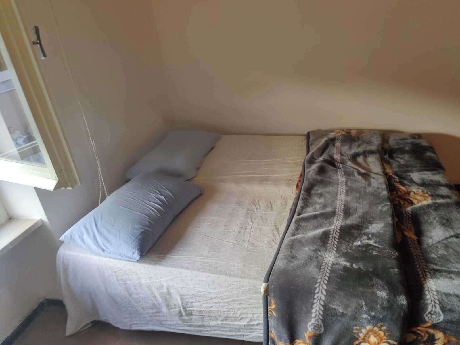 A small, single-bed room with a plain mattress and a grey blanket. There is a window on the left side, and a door on the right.