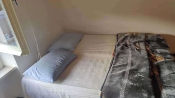 A small, single-bed room with a plain mattress and a grey blanket. There is a window on the left side, and a door on the right.