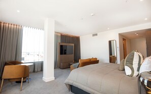A modern hotel room with a large bed, a TV, a seating area, and a view of the city.