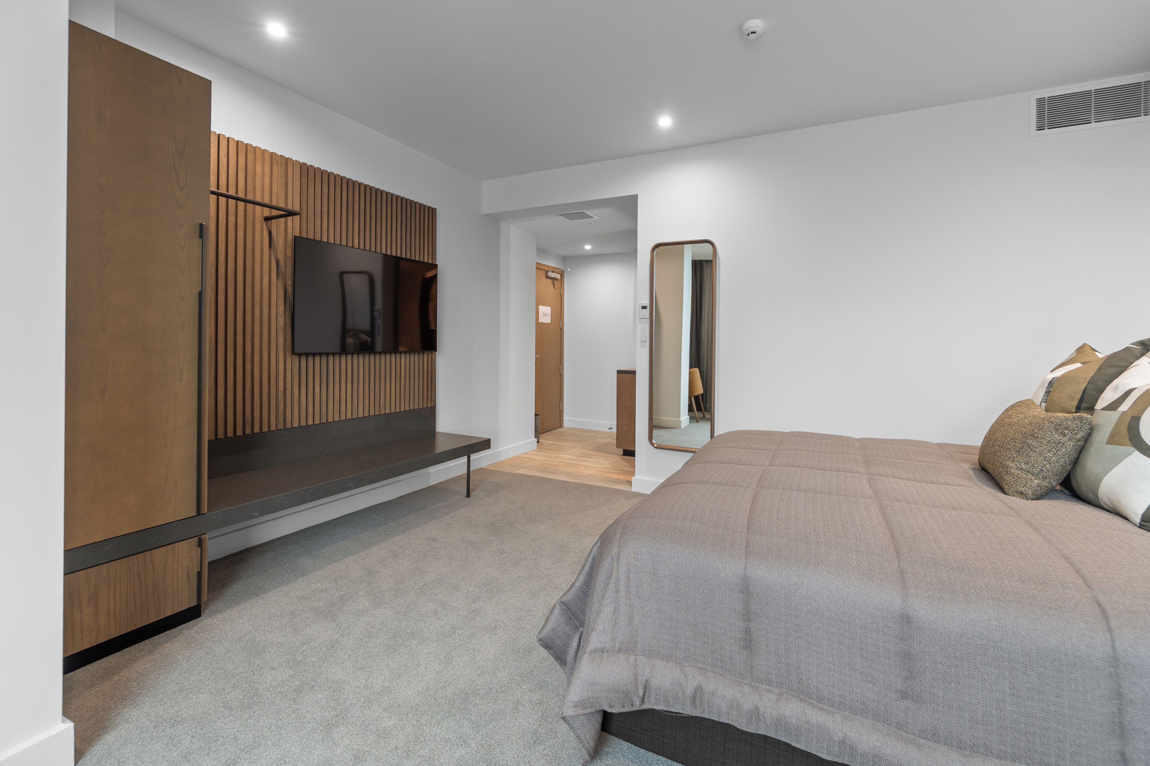 Ein modernes Schlafzimmer mit einem großen Bett, einem an einer Holzpaneelwand montierten Fernseher, einer Bank und einem Spiegel.