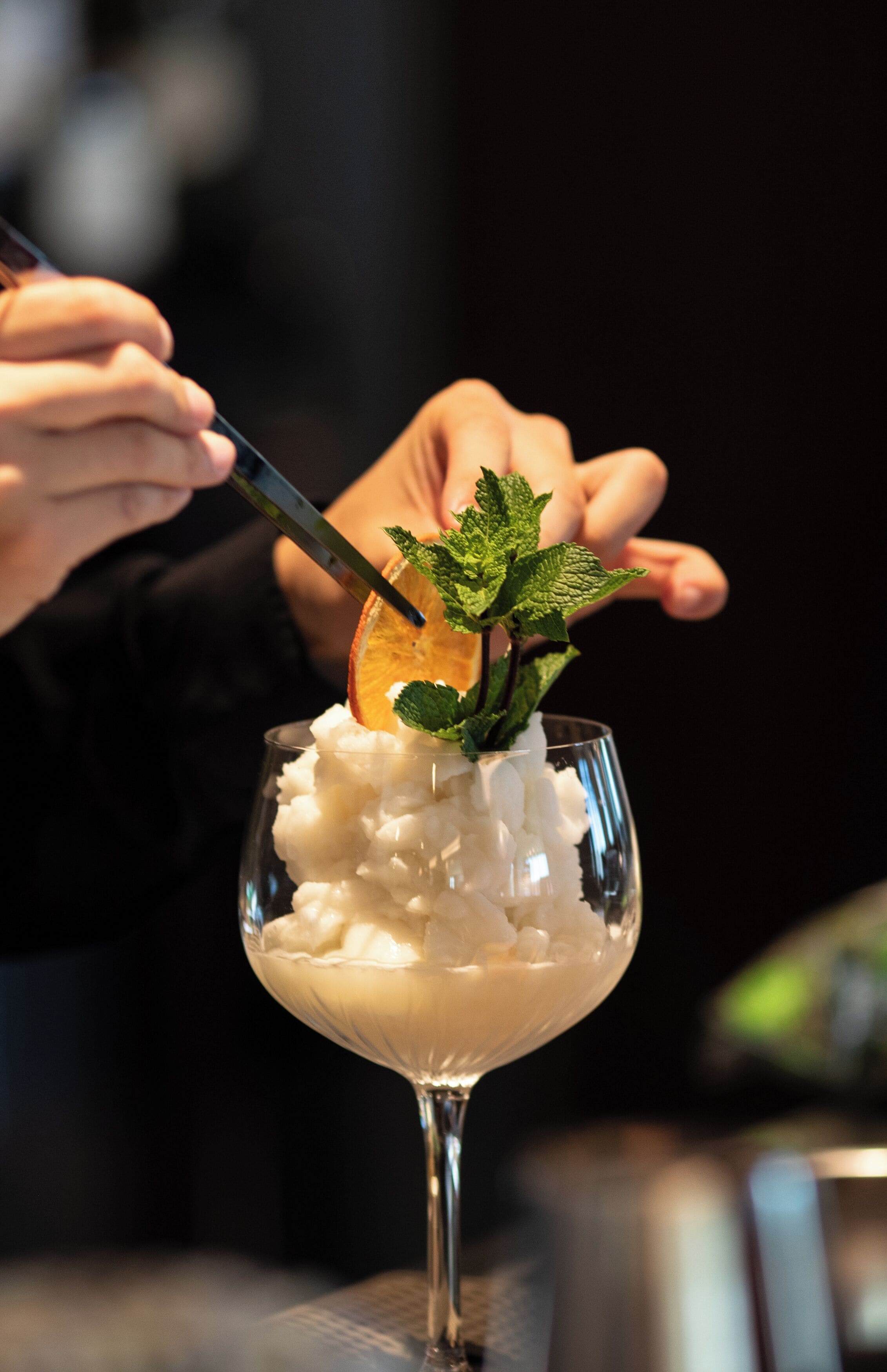 Een cocktail met ijs in een longdrinkglas, versierd met muntblaadjes en een schijfje sinaasappel.