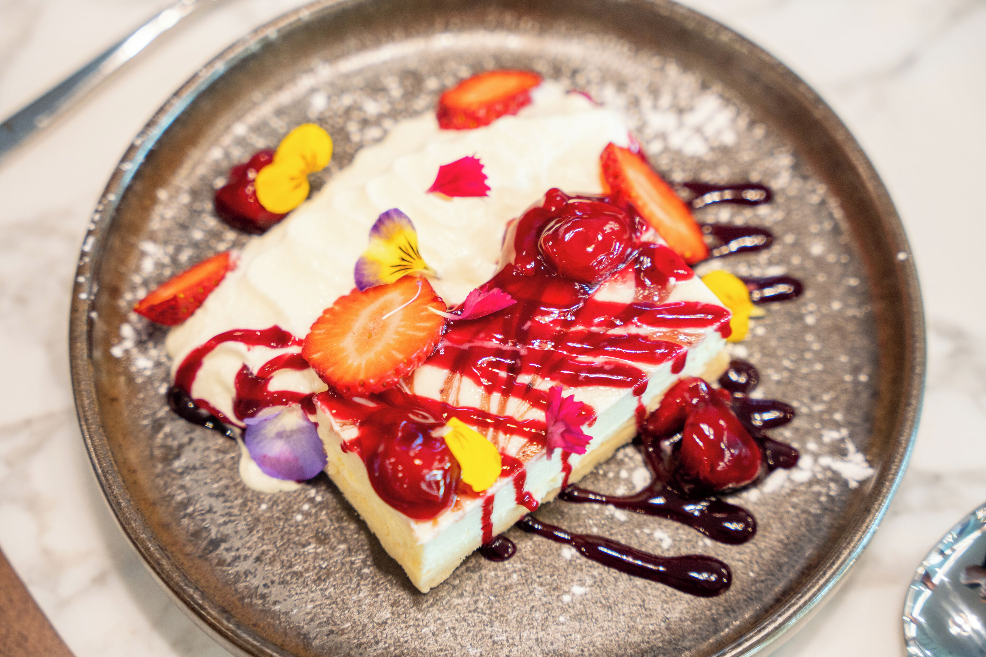 Une assiette de dessert avec une base crémeuse, garnie de fraises, de cerises et d’une sauce rouge, et décorée de fleurs comestibles.