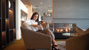 A woman sitting on a sofa in a modern lounge area with a fireplace. - Distinction Invercargill Hotel (Invercargill)