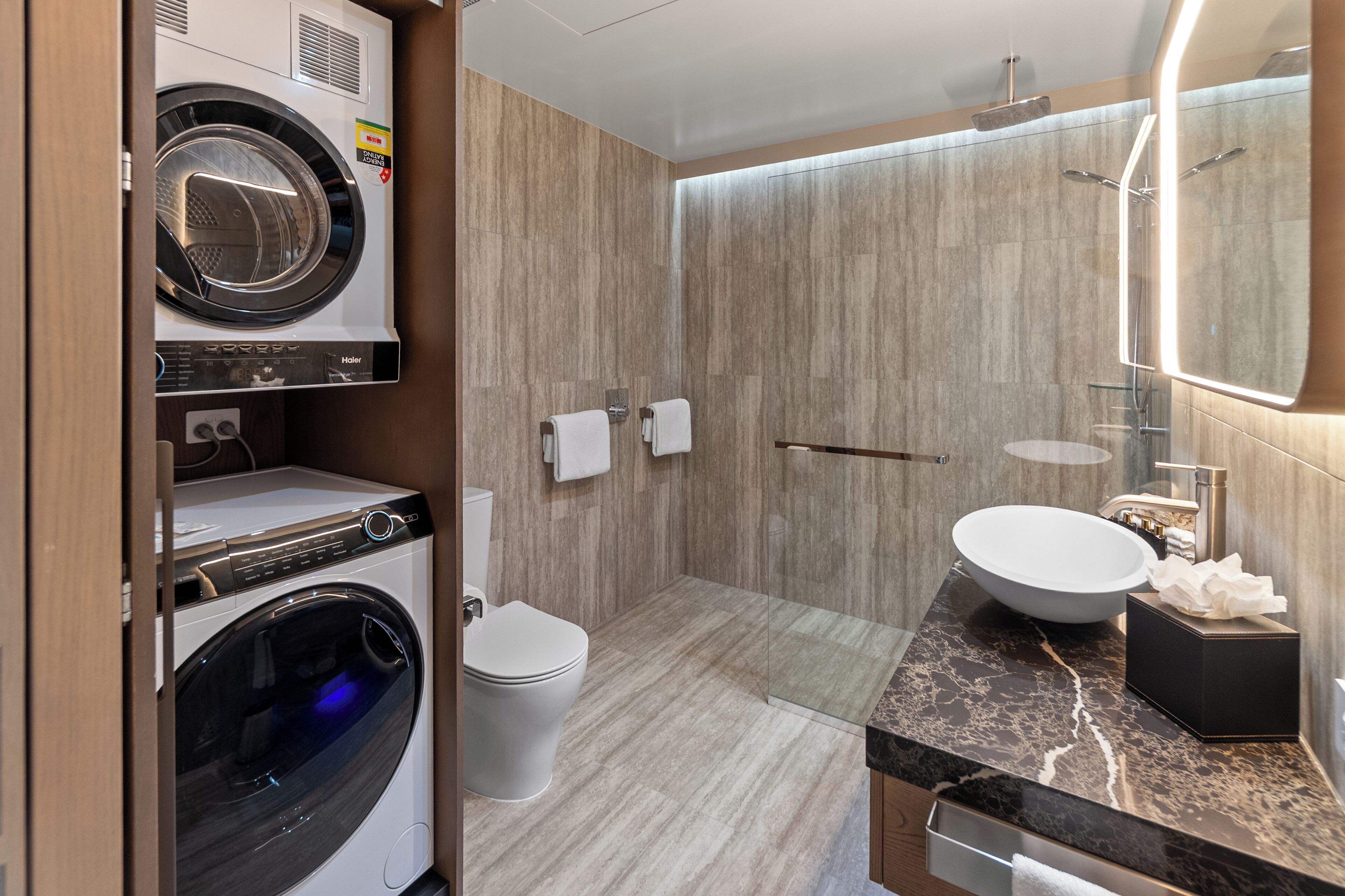Un baño moderno con lavadora y secadora integradas, ducha con mampara de vidrio y encimera de mármol.