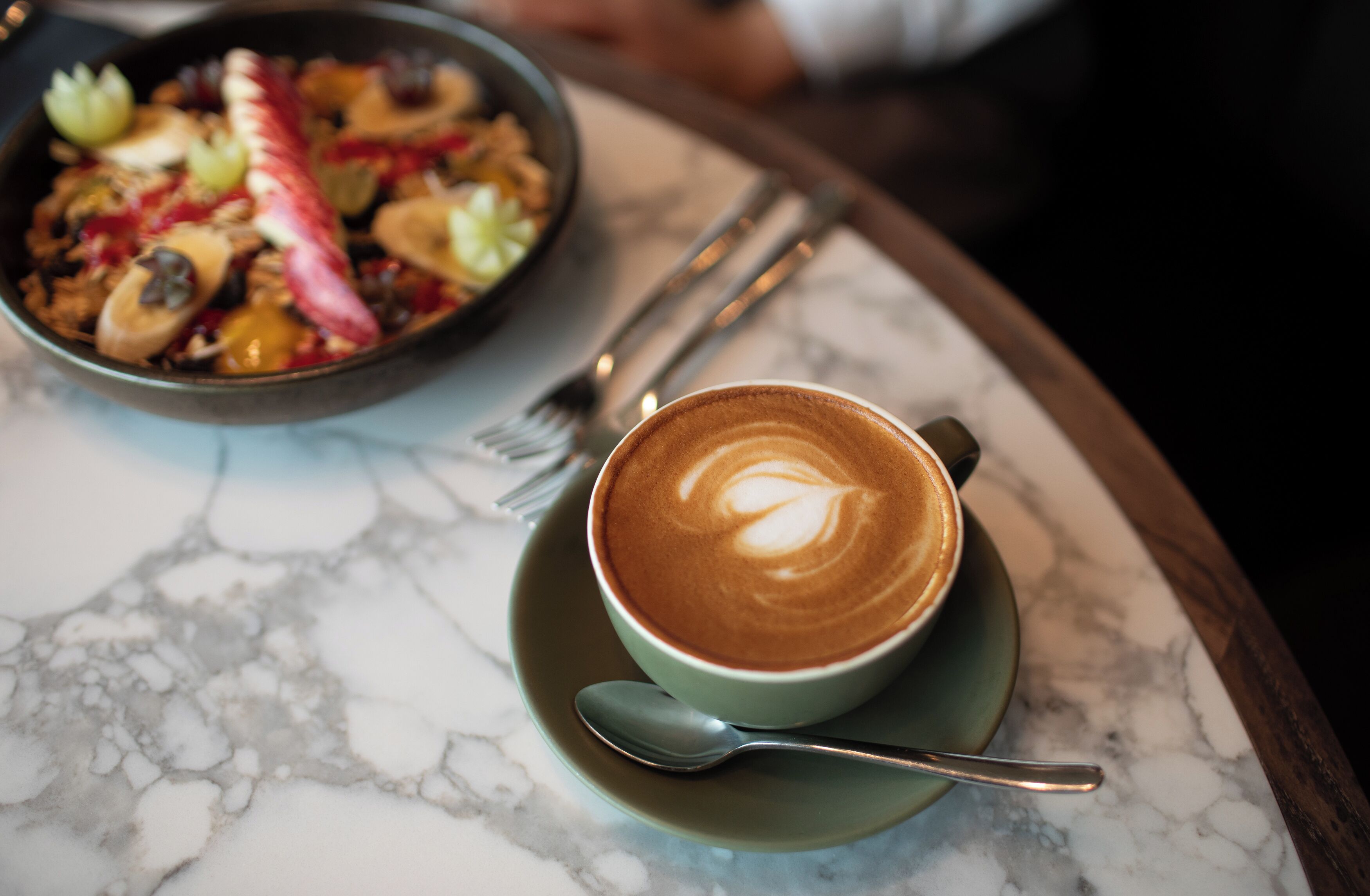A cup of coffee with latte art on a saucer, a spoon, and a fork on a marble table.