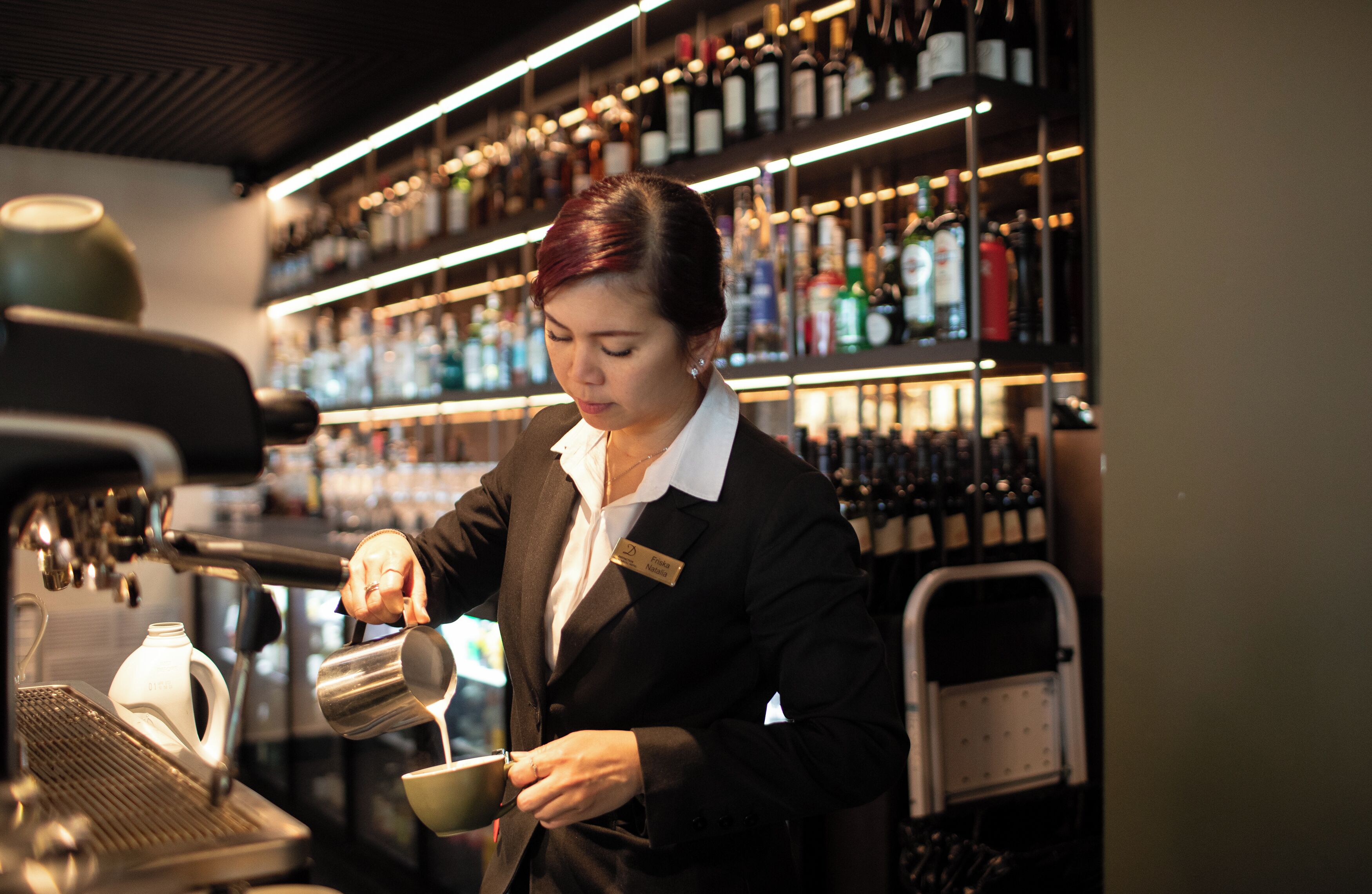 Un barista derrière un appareil à café, une étagère remplie de différentes bouteilles et une cafetière.