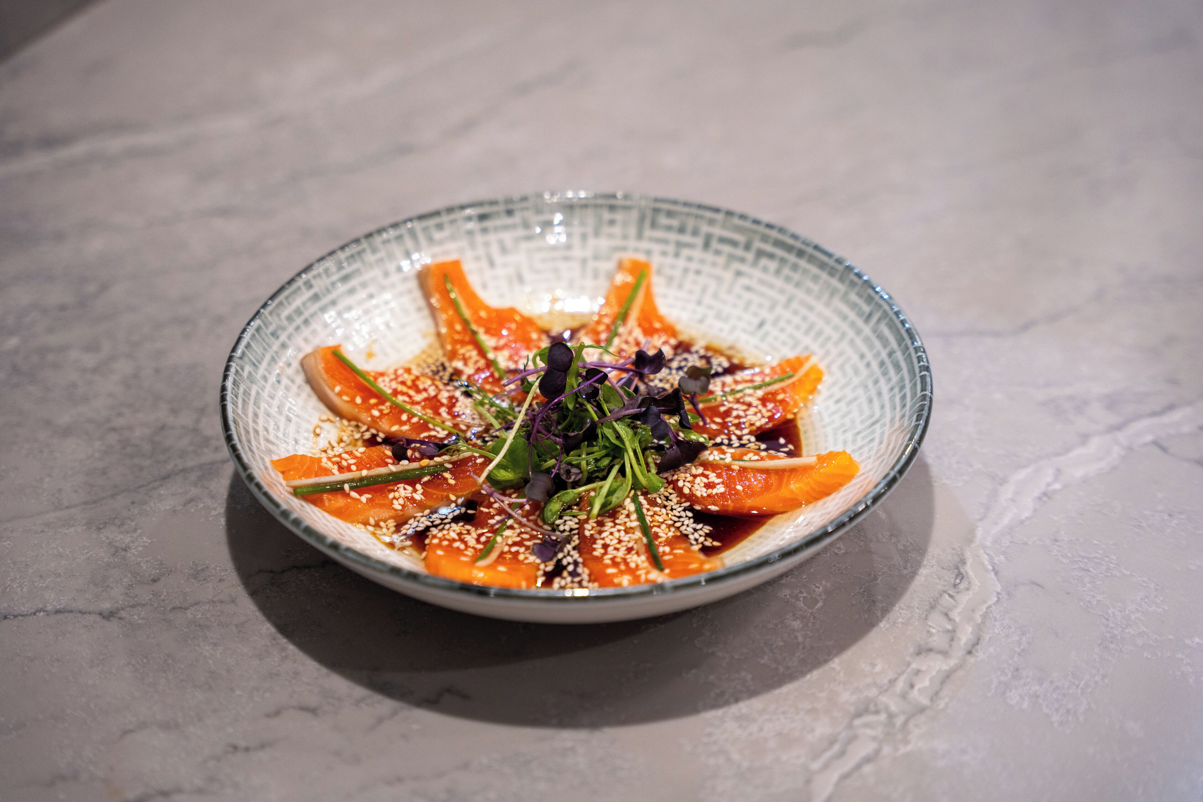 A round glass bowl with a textured rim containing a dish of food, garnished with sesame seeds and greens, on a marble surface.