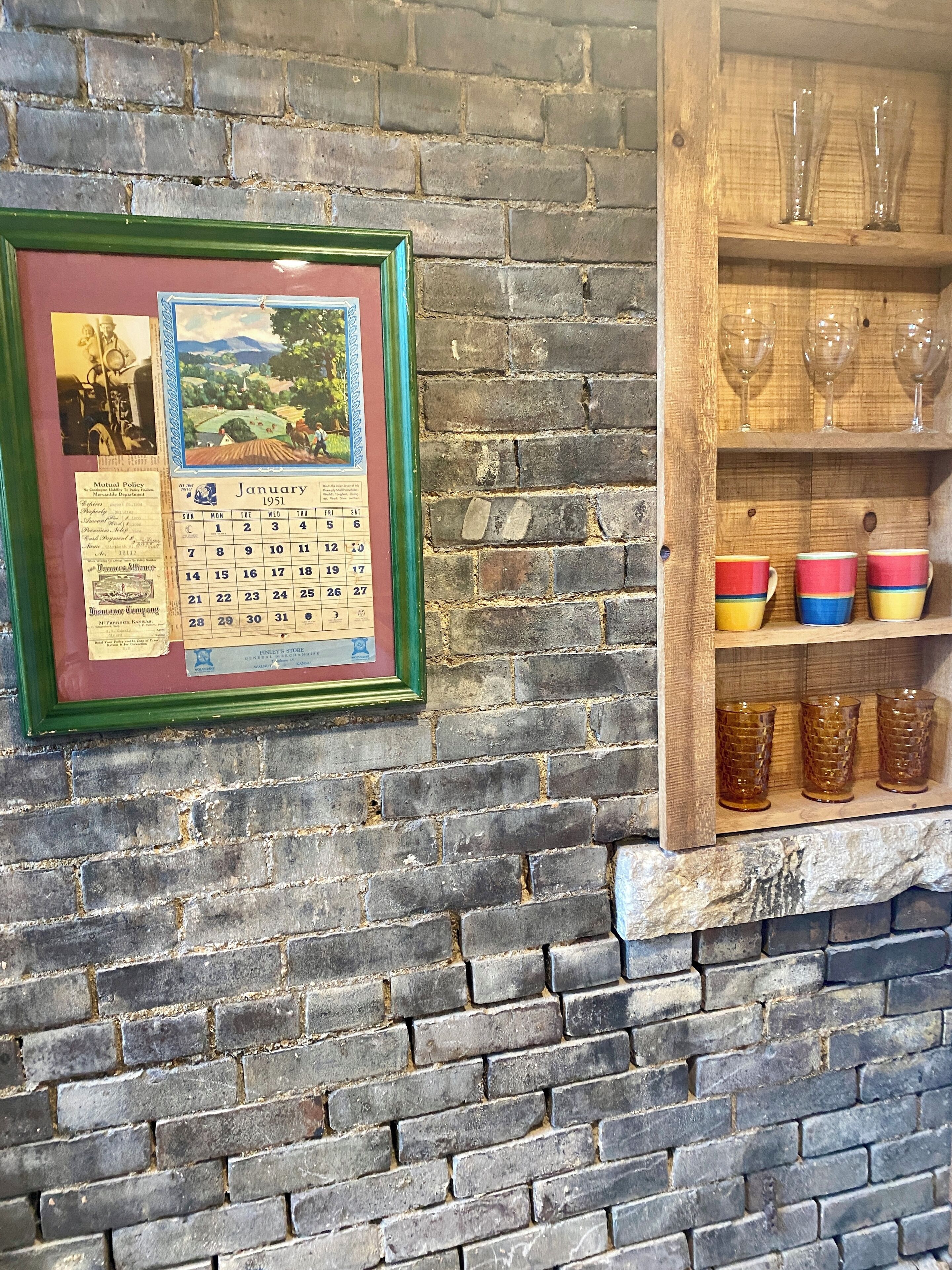 Un mur de briques avec un calendrier encadré et une étagère sur laquelle sont posés des verres et des tasses.