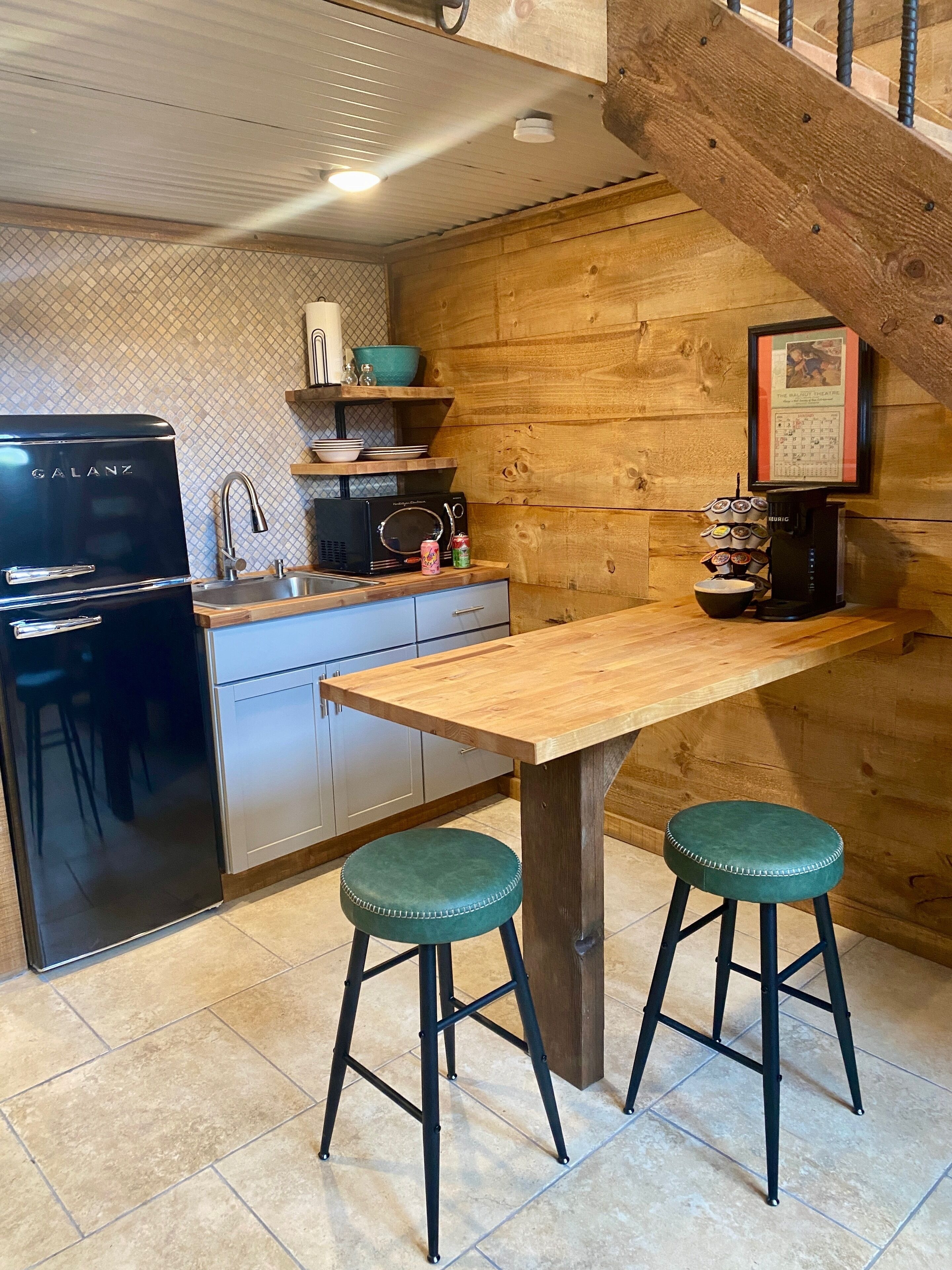 Une cuisine avec une table en bois, deux tabourets, un réfrigérateur, un four à micro-ondes et une machine à café.