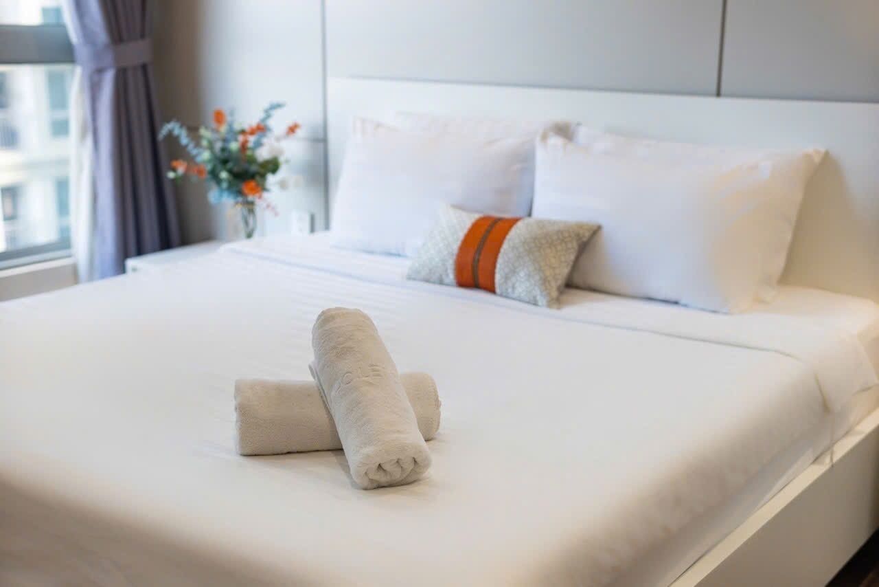 A neatly made bed with white linens and two rolled towels, a vase with flowers, and a window with curtains.