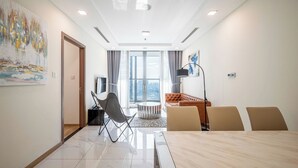 Quarto de hotel moderno com uma janela grande, área de refeições com mesa e cadeiras, sofá e televisão.