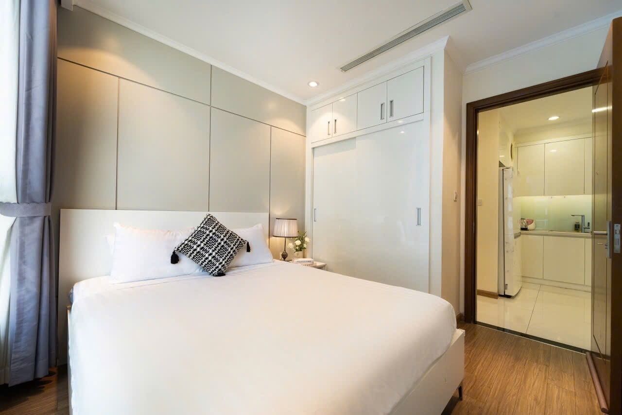 Een hotelkamer met een groot bed, een kledingkast, een nachtkastje met een lamp, en uitzicht op de keuken door een geopende deur.