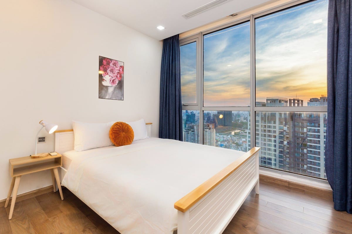 Een moderne hotelkamer met een groot bed, een nachtkastje, een bedlampje, een stadsgezicht door grote ramen en een bloemkunstwerk in een lijst aan de muur.
