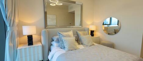 A modern bedroom with a large bed, bedside lamps, a mirror, and a view of the city through the window.