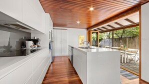 A modern kitchen with white cabinetry, a wooden ceiling, and a view of a backyard with a wooden fence and outdoor seating. - El Cid - Jervis Bay Beach House (Vincentia)