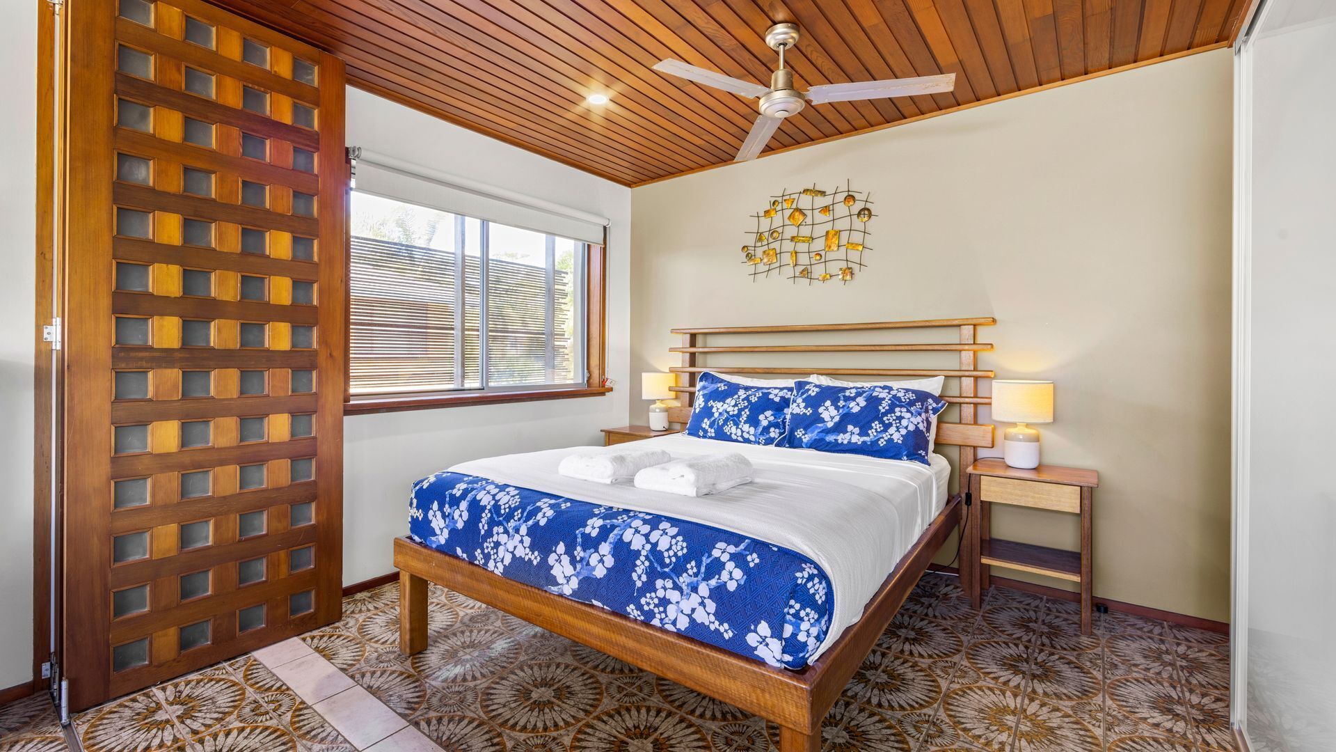 A bedroom with a wooden ceiling, a bed with a floral bedspread, two bedside lamps, and a window with blinds.