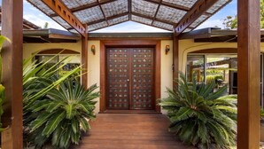A wooden entrance with a glass door, surrounded by large tropical plants. - El Cid - Jervis Bay Beach House (Vincentia)