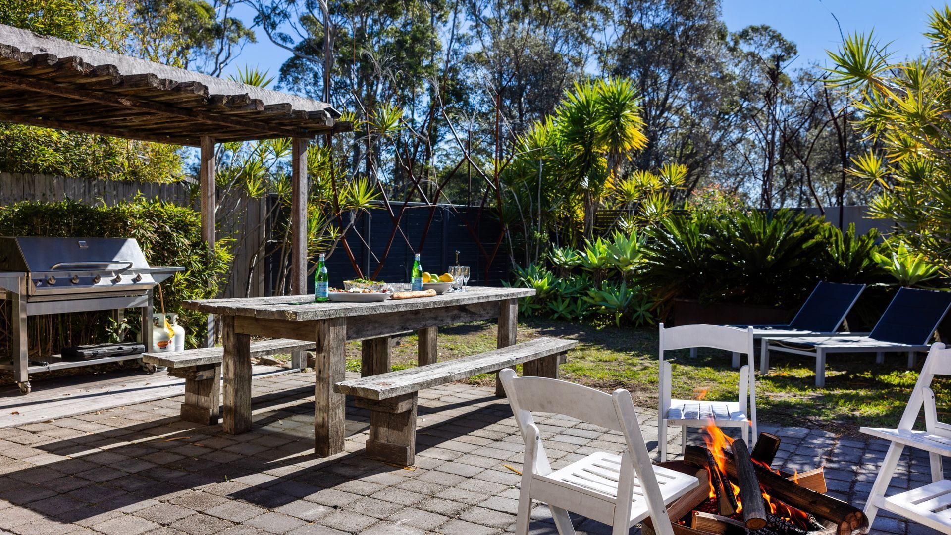 Un patio al aire libre con una parrilla, una mesa con comida y bebidas, y sillones alrededor de una chimenea.