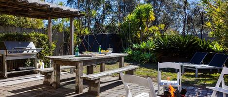 Un patio al aire libre con una parrilla, una mesa con comida y bebidas, y sillones alrededor de una chimenea.