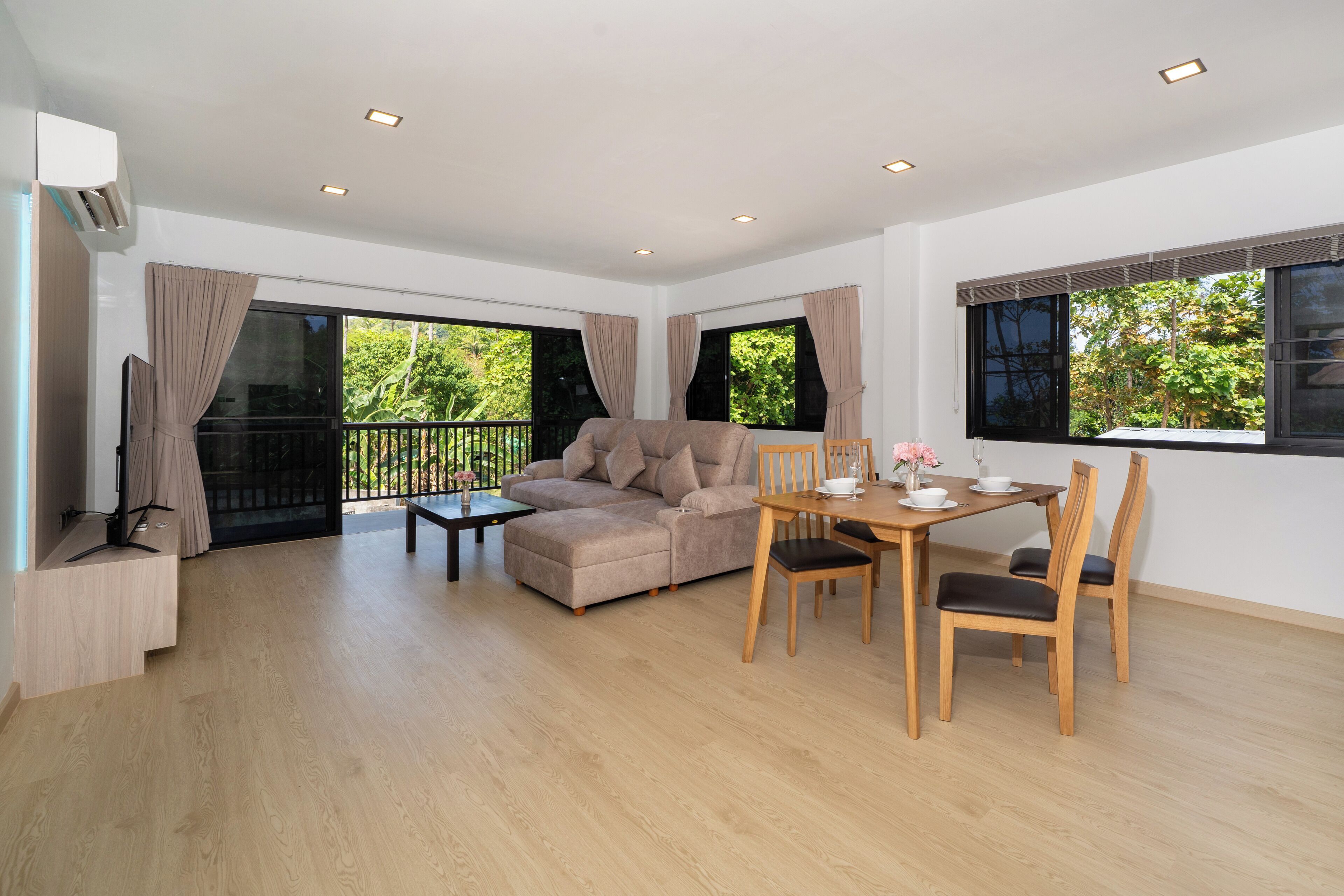 A spacious living area with a beige sofa, a dining table with chairs, and large windows offering a view of greenery.