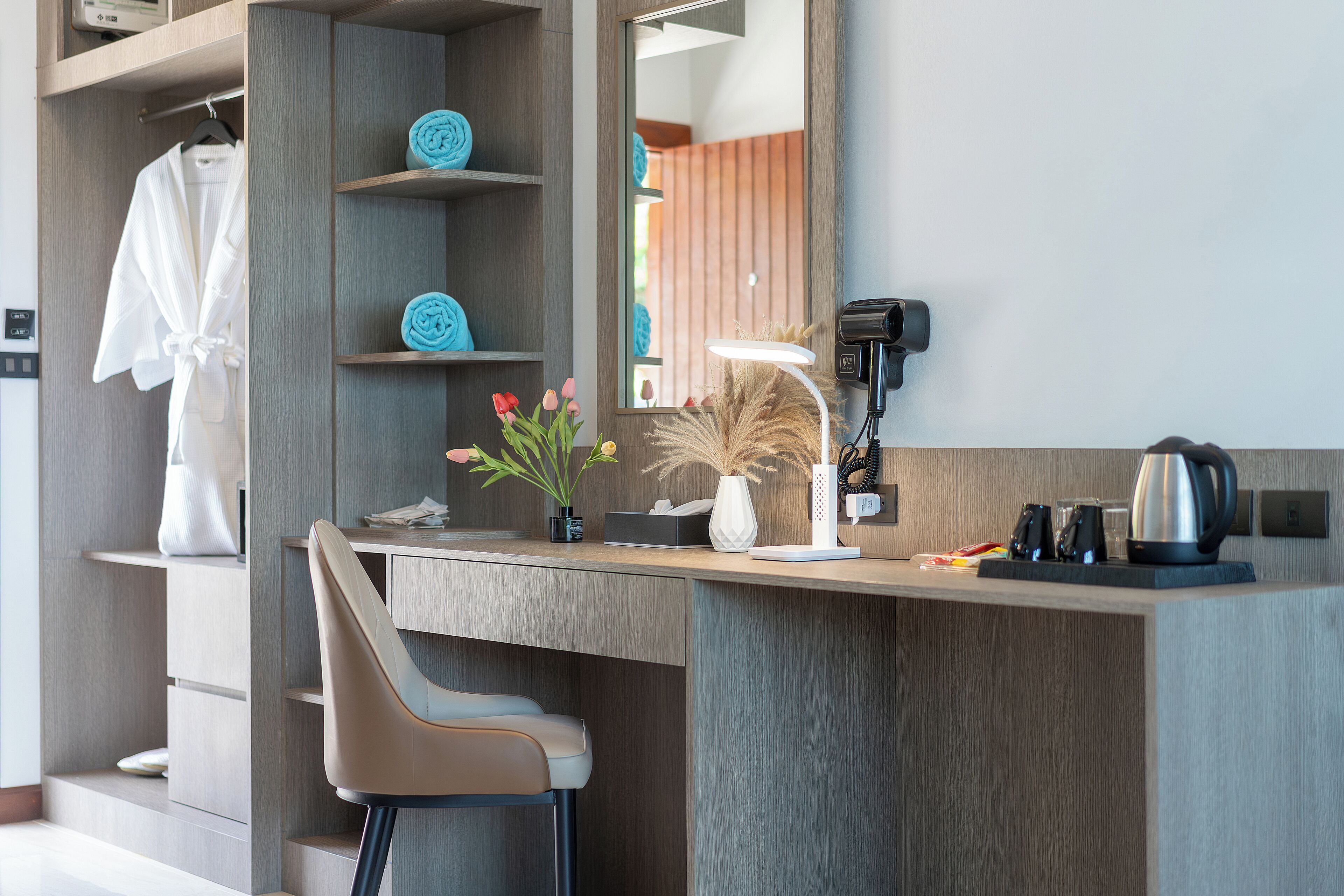 A modern hotel room with a wooden desk, a chair, a robe, towels, a mirror, a lamp, a vase with flowers, and a coffee maker.