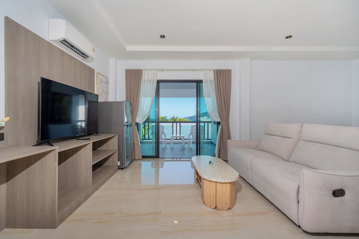 A modern living room with a flat-screen TV, a sofa, a coffee table, and a balcony with a view.