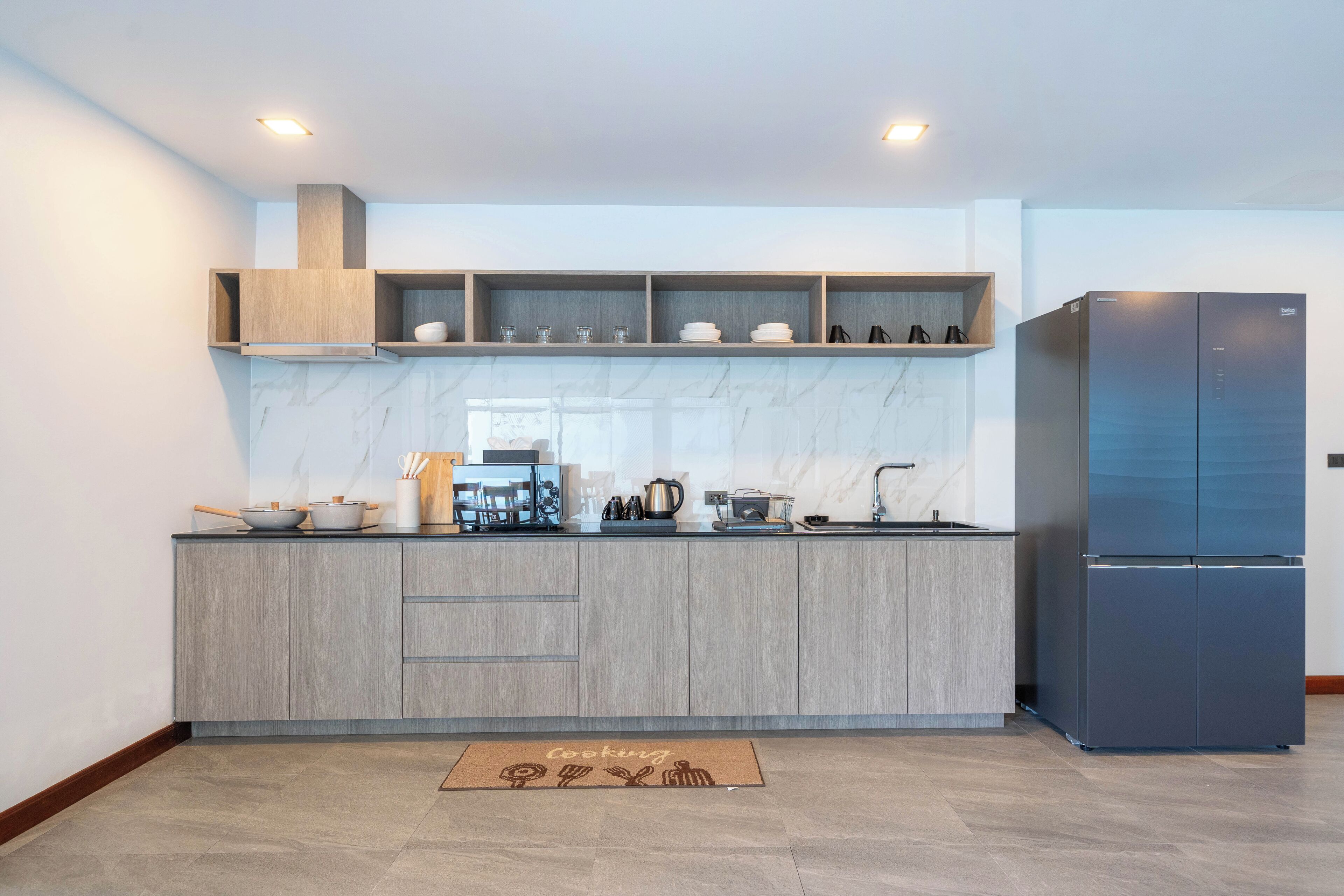 Una cocina moderna con armarios grises, un salpicadero de mármol y un horno empotrado. Se ve un refrigerador negro a la derecha y una estantería de madera sobre la encimera.