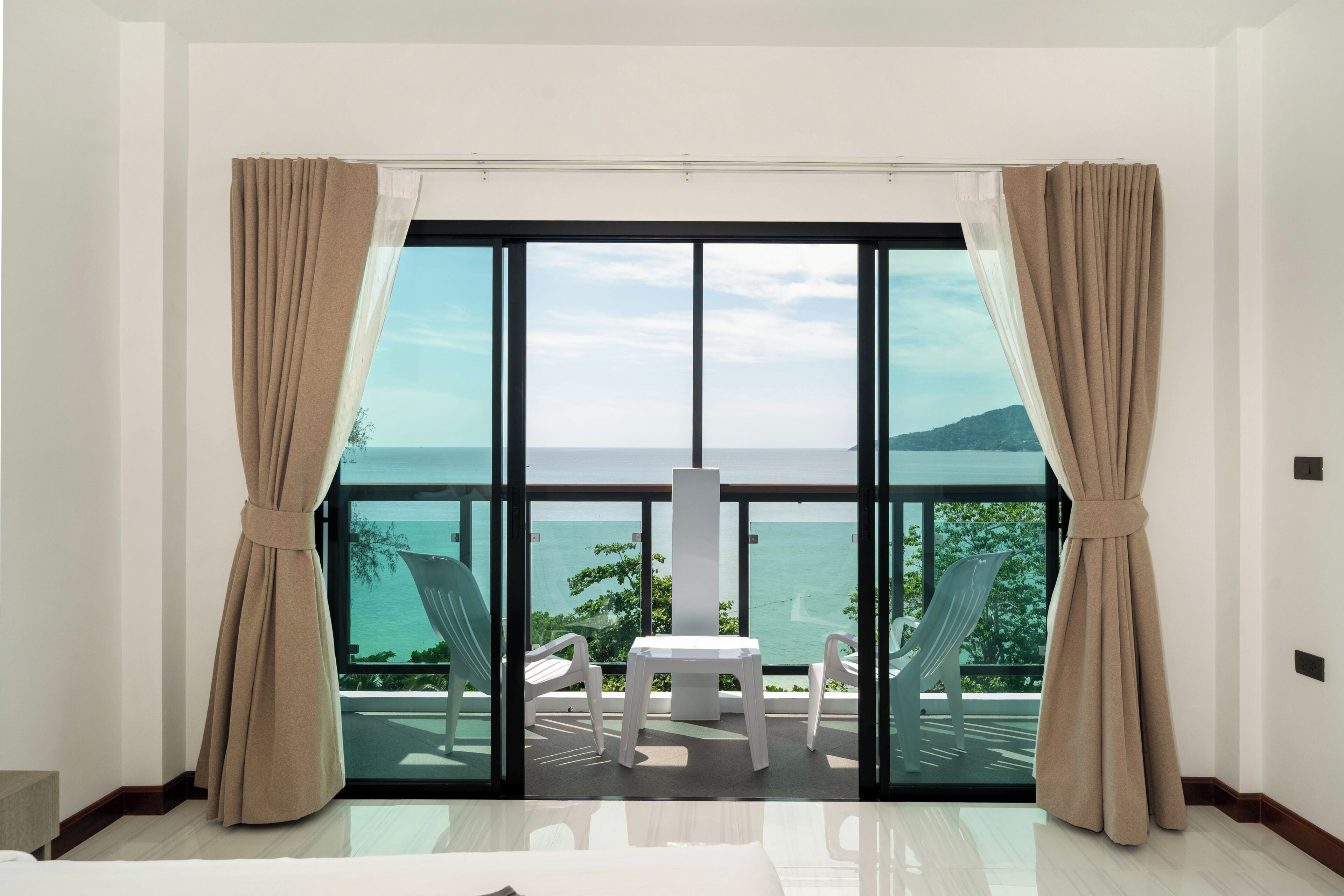 A room with a large sliding glass door leading to a balcony with a view of the sea. There is a small table and two chairs on the balcony.