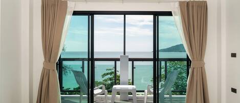 A room with a large sliding glass door leading to a balcony with a view of the sea. There is a small table and two chairs on the balcony.