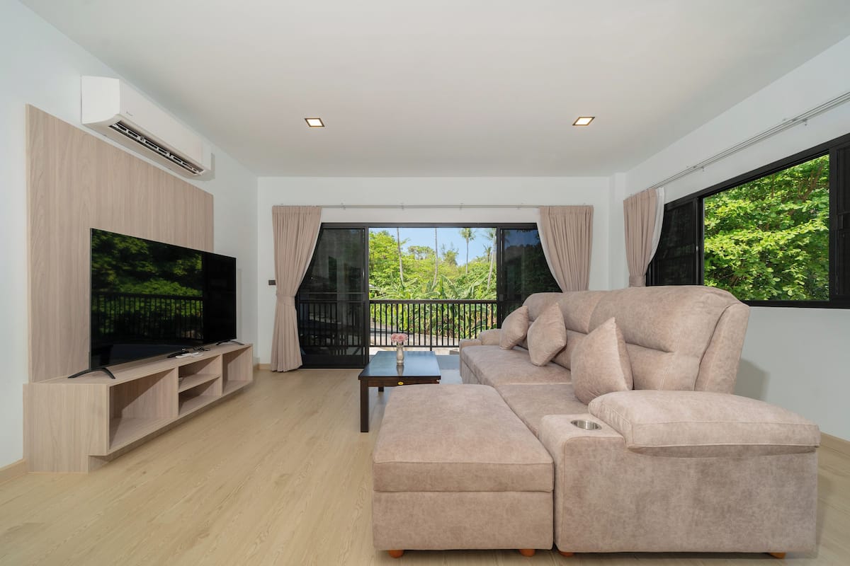 A modern living room with a large flat-screen TV, a beige sectional sofa, a small coffee table, and a balcony with a view of greenery.