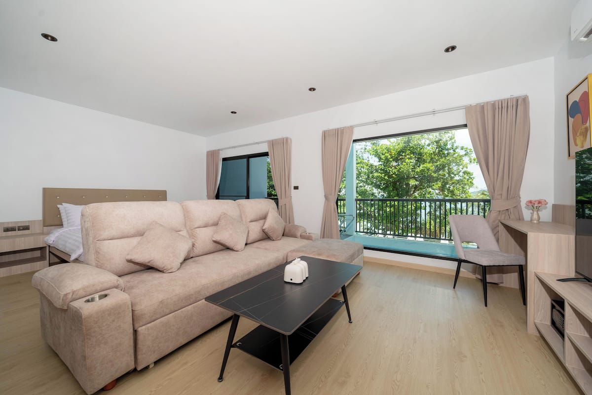 A modern living room with a sofa, a coffee table, and a TV. There is a balcony with a view of trees.