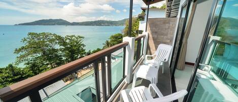Un balcon avec des chaises en plastique blanches, une balustrade en verre et une vue sur la mer et les collines.