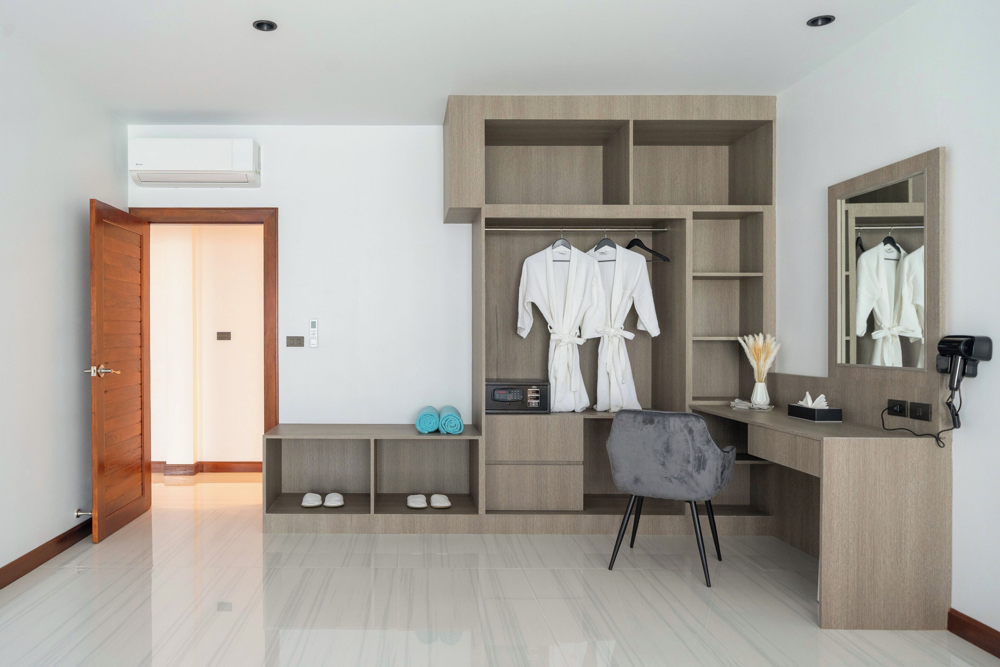 A modern bedroom with a wooden wardrobe, a desk, and a chair. There is a mirror on the wall and a door leading to another room.