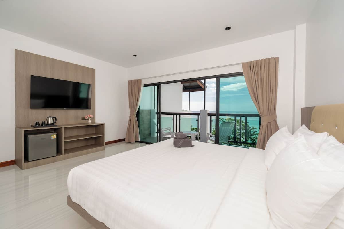 A hotel room with a large bed, a flat-screen TV mounted on the wall, a minibar, and a balcony with a table and chairs.