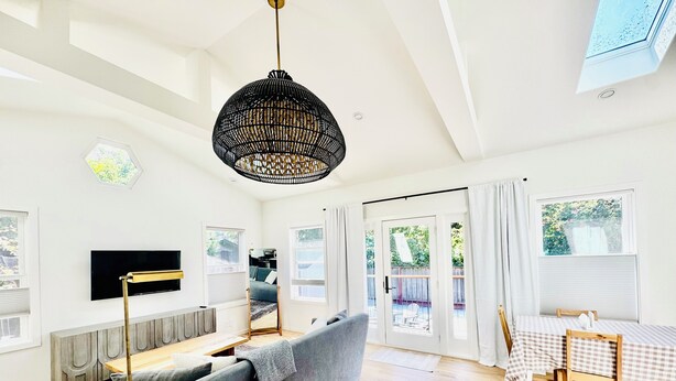 Uma sala de estar moderna com um sofá cinza, piso de madeira, luminária pendente preta, televisão de tela plana e uma área de jantar com toalha de mesa xadrez.