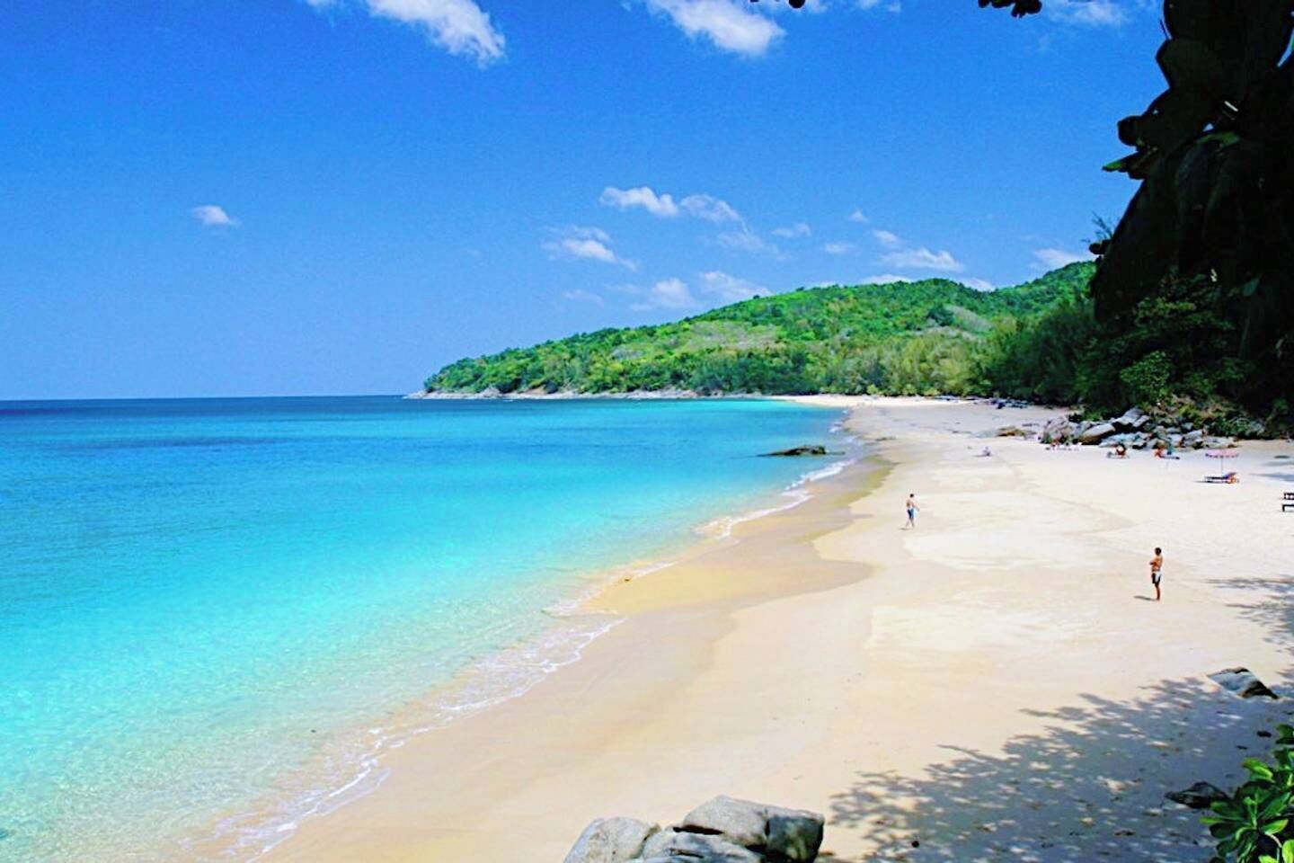 A beach with clear turquoise water, a sandy shore, and a green hill in the background.